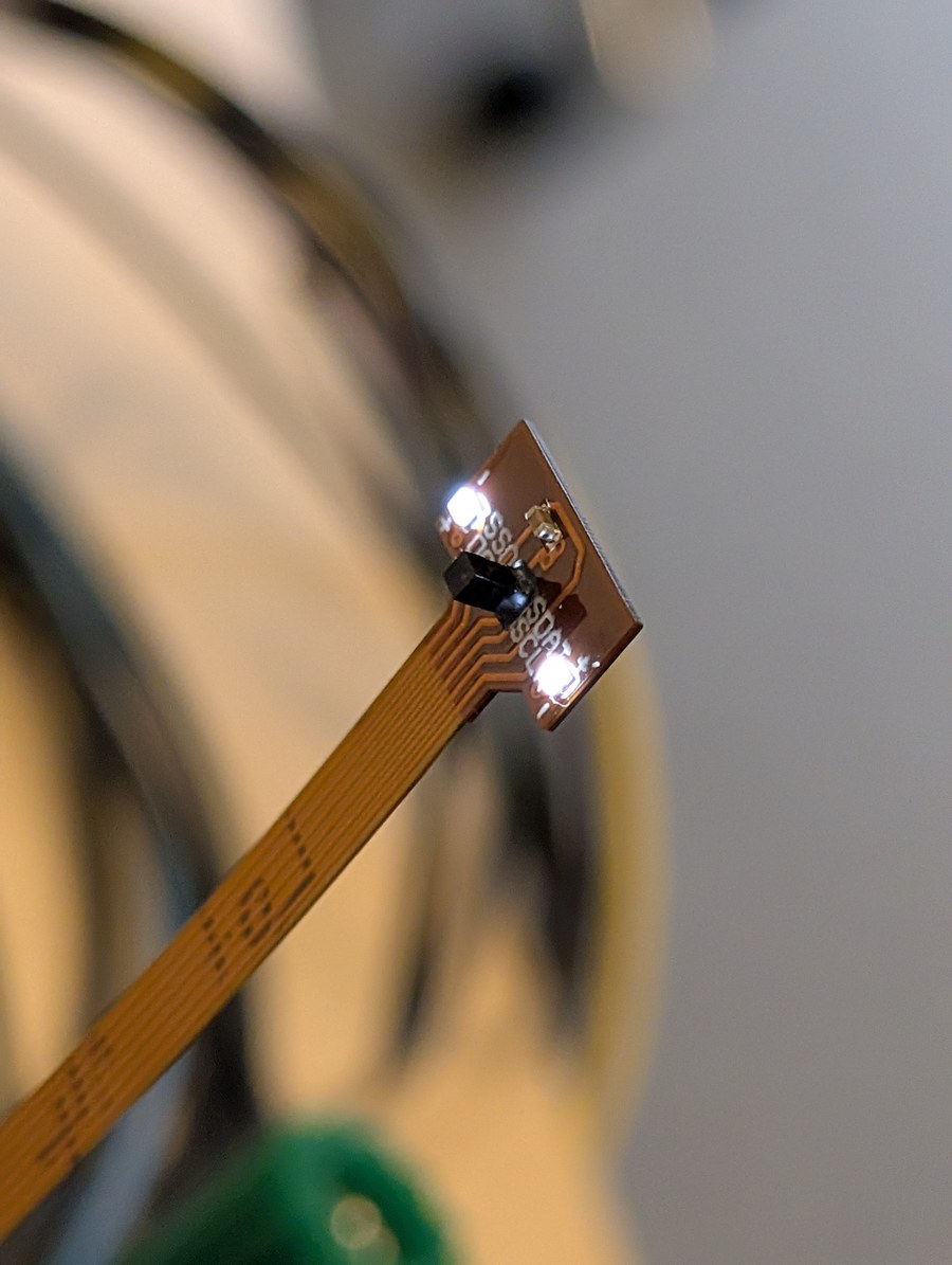 The NanEyeC on the tip of its flex, a black cube about a millimetre across, with a lit white LED either side of it